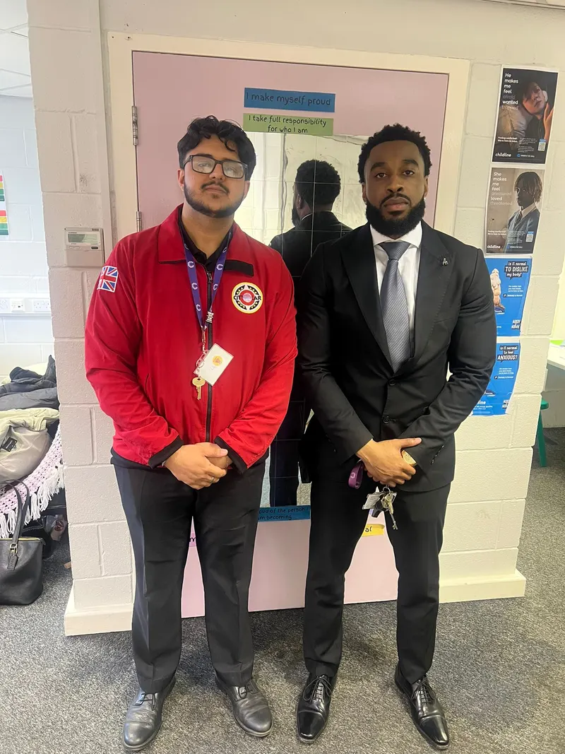 Two mentors standing together in a school corridor, in front of a pupil display board reading “I make myself proud”.