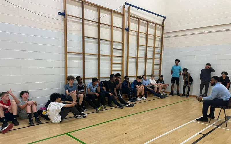 A mentor holding a basketball, seated and talking to a group of pupils sitting along the wall of a school sports hall.